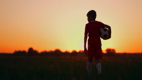 Tracking-the-Boy-goes-to-the-field-at-sunset-with-a-soccer-ball-looking-into-the-distance-and-dreams-of-becoming-a-successful-football-player.-Watch-the-sunset-in-the-field